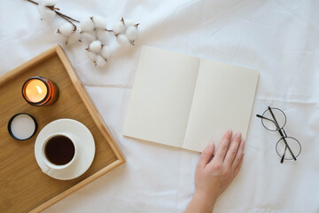 Cozy reading a book in bed. Hand on a book, a tray with a coffee cup, a burning candle and glasses on a white sheet