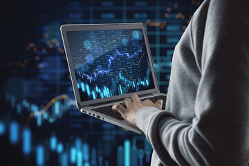Close up of hand holding and using laptop with glowing forex index chart/graph on dark background....