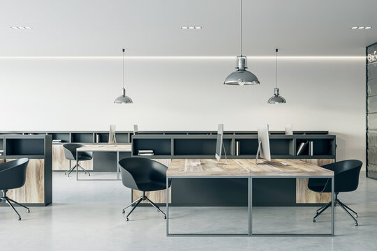 Side view on wooden work table with modern computers and black chairs on glossy concrete floor in coworking office with light wall background and stylish lamps hanging from ceiling. 3D rendering