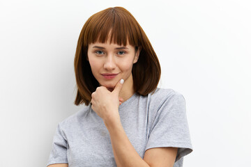 Fototapeta premium a sweet, thoughtful, beautiful woman stands on a white background in a gray cotton T-shirt and thoughtfully holds her hand near her face