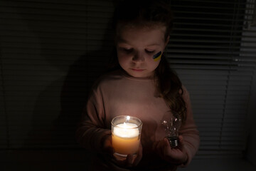 A girl with a painted flag of Ukraine on her cheek in a dark room holds a candle and an electric...