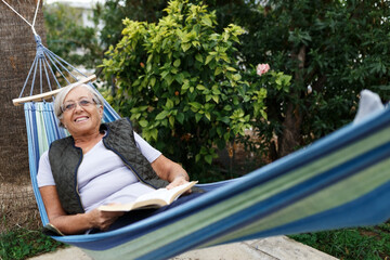 Smiling senior good-looking grey hair woman wearing glasses while reading in hammock in the summer garden