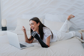 Cheerful young brunette girl with bionic artificial hand laying on bed with laptop makes video call...