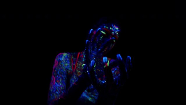 Young female performer with fluorescent multicolor drips on his skin moving hands in front of her own face in dance in uv light on a black background. Painted woman dancer performs in the dark