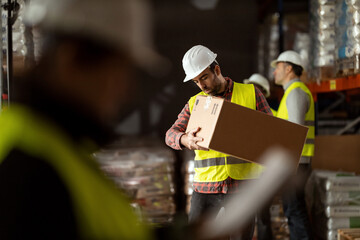 A warehouse worker wearing protective clothing is carrying a box in a large warehouse.