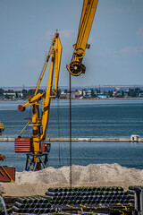 Two port cranes stand against the background of blue water and a blue sky with metal pipes and...