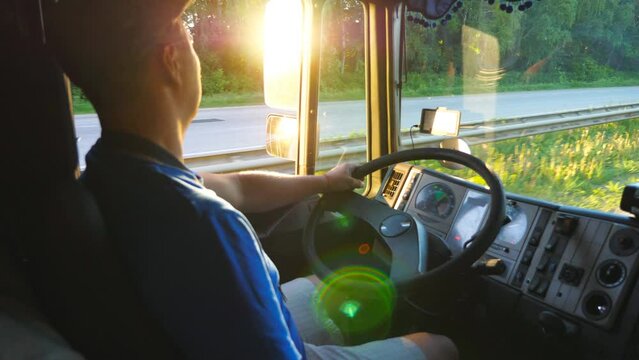 Close up of lorry driver riding through countryside. Man in hat controlling his truck and carefully watching traffic. Bright sunset light reflecting in side mirror of car. Slow motion