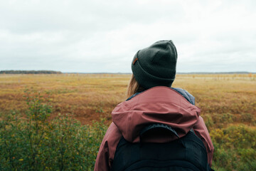 Backview of blonde girl in front of a plain. Young hiker in green hat observes the landscape. Adventure and travel in cold northern European countries. Solitude and thoughts. Traveling alone.