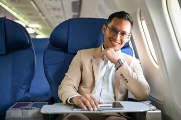 Handsome Asian businessman sits at the window seat in business class, hand on chin, smiling