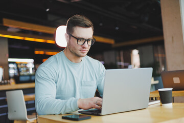 A young promising man in casual clothes sits at a table and works on a laptop. Work space for remote work.