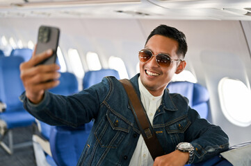 Happy Asian male travel blogger or tourist taking selfie by his smartphone during the flight.