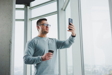 Naklejka na ściany i meble Smiling man in casual clothes standing near window with coffee and looking at smartphone in modern workspace with big window during daytime