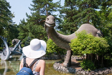 Novosibirsk, Russia, August 2022: A girl in a white hat looks at a dinosaur near a lake in the...