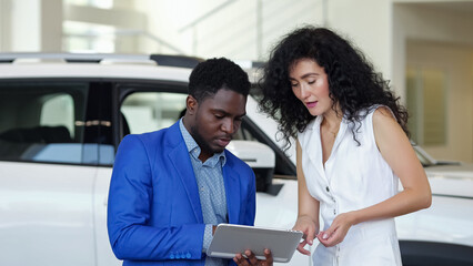 African American male car dealer presents car to female customer in automobile salon. Black...