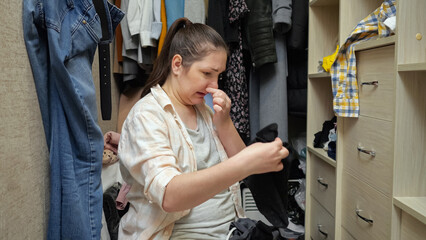 Brown-haired woman sorts dirty male clothes in walk-in closet and looks with upset and disappointed expression. Lady smells stale clothes and feels disgusted - obrazy, fototapety, plakaty