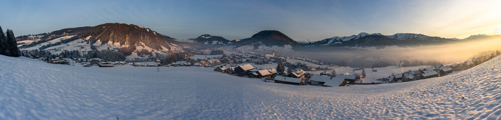 Panorama mit Waldrand, Dorf und Schnee auf Wiese und im Hintergrund die Berge des Bregenzerwaldes. verschneite Landschaft, Nachmittagssonne vor Sonnenuntergang, beleuchtete Landschaft im Winter, 