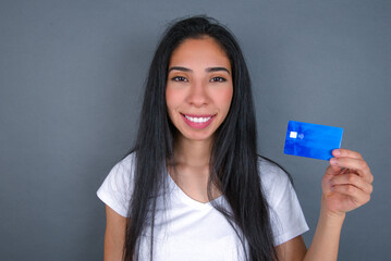 Young beautiful hispanic woman wearing white t-shirt over gray studio background, smiling and presenting a blue credit card. Online shopping and finances concept. 