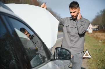 A young man near a broken car with an open hood on the roadside.