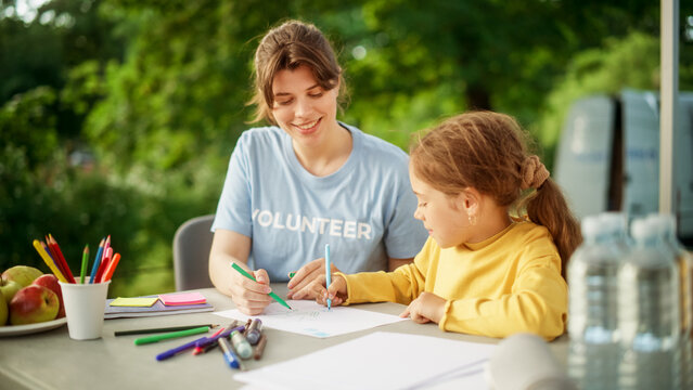Talented Small Girl in a Yellow Jumper Drawing Beautiful Pictures Together with a Female Volunteer from a Local Charity Organization. Humanitarian Aid Worker Helping Child with Arts Class Homework.