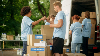 Positive Group of Young Adult Volunteers Loading Food Rations and Humanitarian Donations for Poor People in Need into a Van. Charity Worker Using Tablet Computer, Keeping Inventory Documents in Order.