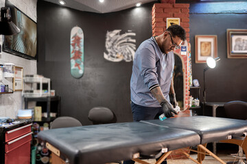 A tattoo artist is cleaning the studio stretcher with a spray