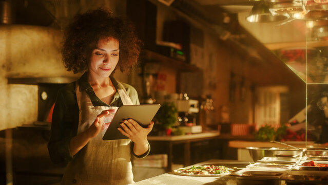 In Restaurant: Beautiful Female Chef Using Laptop Computer. Authentic Italian Pizzeria With Delicious Organic Eco Food. Bi-racial Female Entrepreneur Working in Her Small Business Family Shop - Powered by Adobe