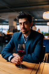 Vertical portrait of confident elegant man in fashion suit holding glasses of red wine sitting at table in restaurant with dark interior, looking at camera. Bearded male resting, having dinner alone.