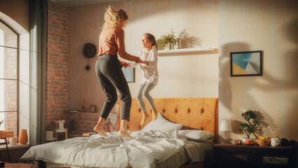 Adorable Little Girl Having Fun at Home with Young Beautiful Mother. Family Playing at Home, Cheerfully Jumping on Bed in Stylish Loft Apartment with Big Window. Happy Childhood.