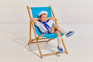 Portrait of little boy, child in image of seaman in vest posing, sitting on chair over grey background. Relaxation, cruise