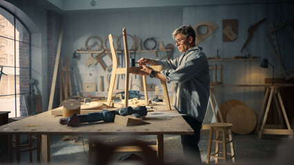 Portrait of a Carpenter Putting on Glasses, Looking at a Blueprint and Starting to Assemble a Wooden Chair. Professional Furniture Designer Working in a Studio in Loft Space with Tools on the Walls.