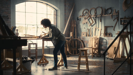 Multiethnic Artisan Woodworker Marking Out Dimensions on a Blueprint Before Creating a Furniture Piece. Black Female Carpenter Working in a Studio in Loft Space with Tools on the Walls.