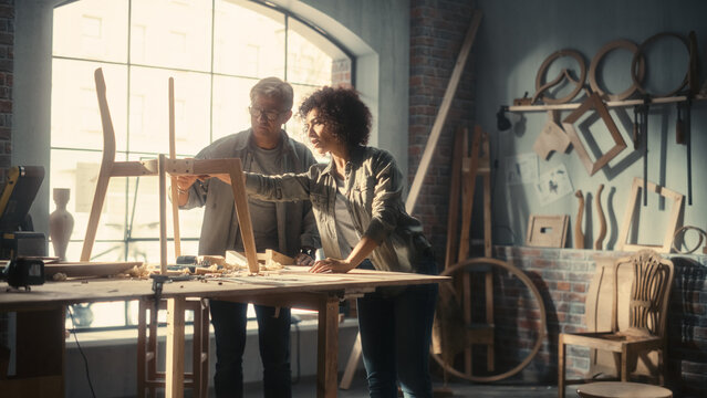 Portrait of Two Carpenters Working Together in a Woodwork Workshop. Multicultural Man and Female Colleagues Looking at a Blueprint on Paper and Discussing a New Chair Design.