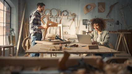 Adult Carpenter and a Young Multiethnic Female Apprentice Working in Loft Studio on a New Chair Design. Small Business Owners Talking About Work, Using Laptop Computer in Stylish Furniture Workshop.