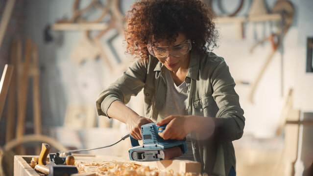 Multiethnic Woman Carpenter Wearing Protective Safety Glasses and Using Electric Belt Sander to Grind a Wood Block. Artist or Furniture Designer Working on a Product Idea in a Workshop.