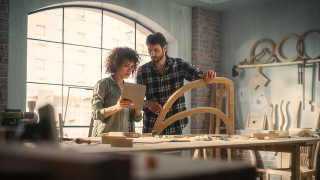 Portrait of Two Small Business Owners Using Tablet Computer and Discussing the Design of a New Wooden Chair in a Furniture Workshop. Carpenter and a Young Female Apprentice Working in Loft Studio. - Powered by Adobe