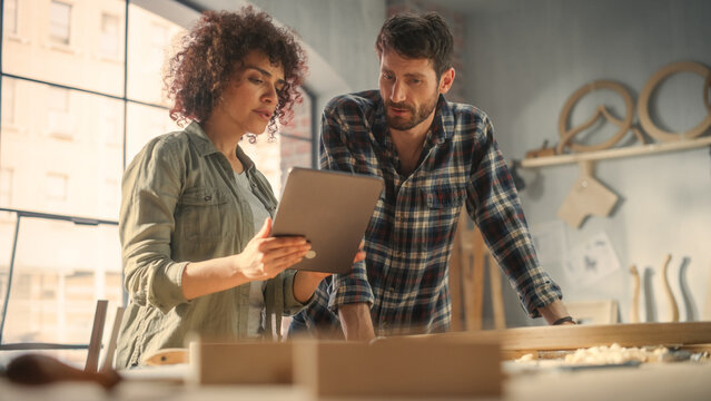 Two Talented Small Business Owners Using Tablet Computer and Discussing the Design of a New Wooden Chair in a Furniture Workshop. Carpenter and a Young Female Apprentice Working in Loft Studio.