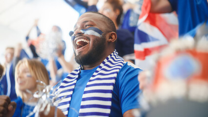Sport Stadium Big Event: Handsome Black Man Cheering. Crowd of Fans with Painted Faces Cheer, Shout for the Blue Soccer Team to Win. People Celebrate Scoring a Goal, Championship Victory