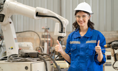 Portrait of female industrial engineer happy working with robotic technology machine. Beautiful woman worker standing in modern metalwork factory at robot welding machine thumb up and smiling.