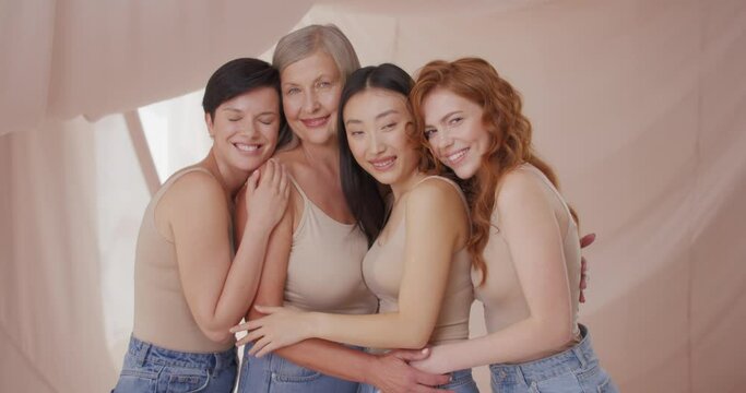 Portrait of four beautiful charming natural women posing smiling hugging wearing nude makeup. Pretty young and mature asian caucasian and ginger female models on beige background. Nude makeup concept.