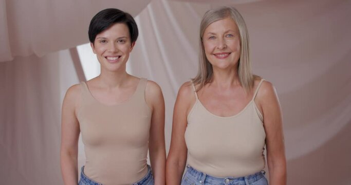 Portait of two beautiful pretty natural women smiling wearing nude light makeup. Young brunette and mature blonde females posing wearing beige tanks in studio. Natural beauty concept.