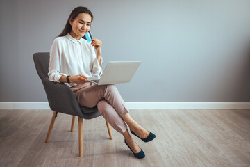 Online shopping, e-commerce, remote banking concept. Lovely young woman sitting in armchair with laptop, using credit card to buy things on web, purchasing in internet store.