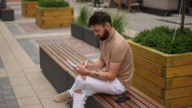Thoughtful bearded man with tattooed hands writing notes in paper notepad sitting on bench in city street in summer day. Creative male artist painting in sketchbook with pen sitting on bench.