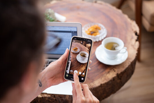 A white woman taking a picture of her cup of tea and french traditional financier dessert using a smartphone camera next to a rustic wooden table with a modern laptop computer on it inside a house