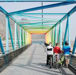 three children of boys with bicycles on a sunny day stand with their backs at the beginning of a multi-colored bridge, the sea and mountains in the distance