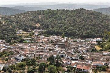 View from above onto the quaint white village of Alájar, Spain, with the rolling hills of the Sierra de Aracena and a hazy autumn sky in the background
