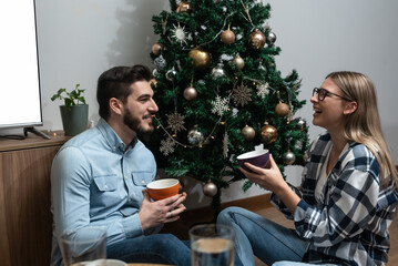 Obraz na płótnie Canvas Young couple sitting on the floor of home in front of Christmas Tree with New Year decoration drinking coffee or tea and enjoying and talking. Woman and man sitting joking and laughing on holiday.