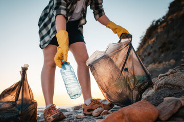 Global environmental pollution. Woman volunteer holding a polyethylene bag and picks up a plastic bottle on the beach. Close up and low angle view. The concept of cleaning the coastal zone