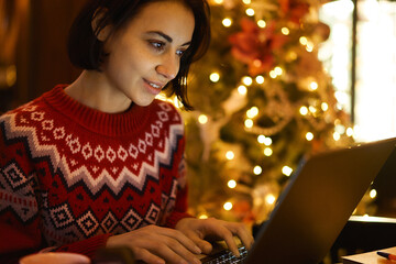 Caucasian female blogger in red winter sweater smiling indoors while texting and reading connected to 4g wireless in coffee shop xmass interior