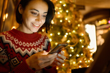 Closeup of pretty smiling woman using smartphone in cafe with christmas festive tree and lights....