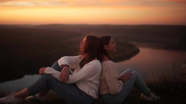 Two teenage girls sit on top of a rock and enjoy the summer sunset. Sisters enjoy youth and freedom. A gust of wind blows the hair of two friends